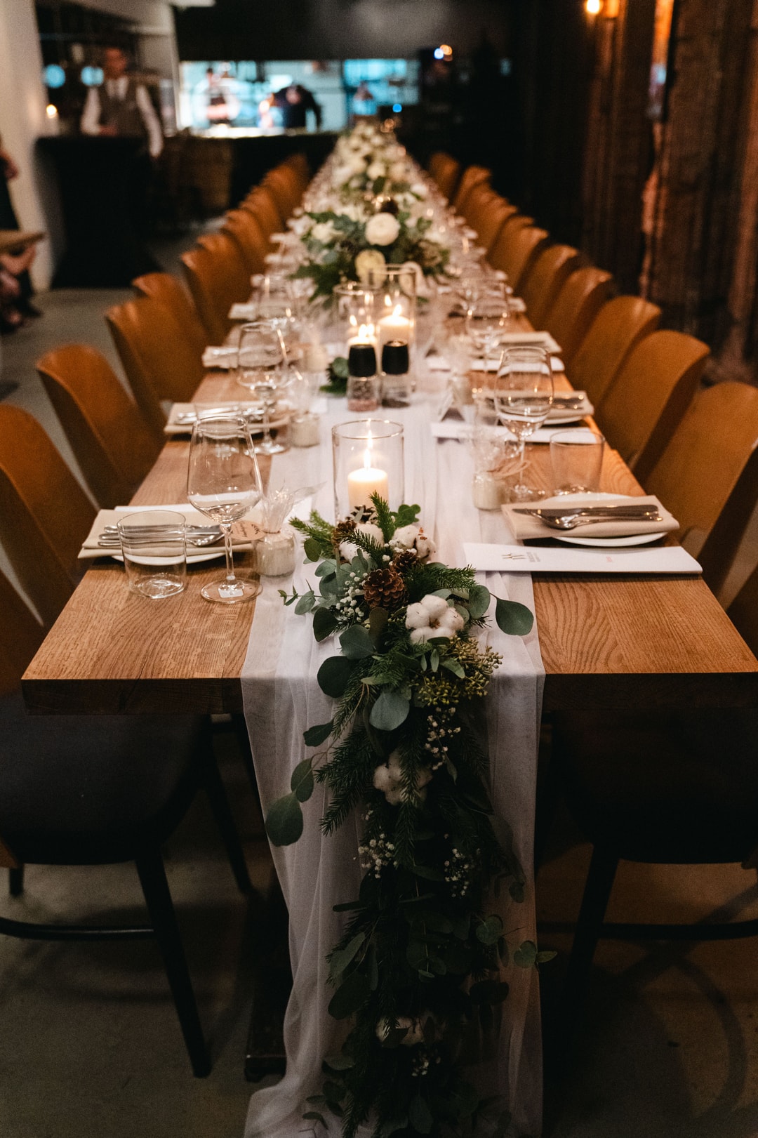 Długi stół weselny z drewnianym blatem, brązowymi krzesłami, białym obrusem, dekoracje sali weselnej z białymi kwiatami i zielenią, świeczniki
