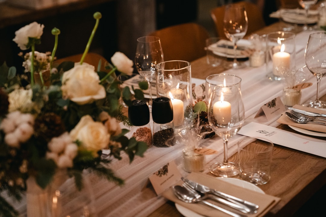 Aranżacja stołu weselnego z białymi kwiatami, świecami w szklanych lampionach, kieliszkami i złotymi sztućcami na jasnym obrusie