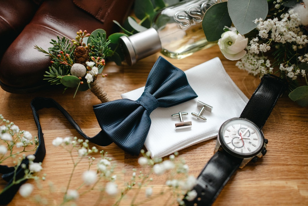 Drewniana powierzchnia z dekoracją stołu: granatowa mucha, białe chusteczki, srebrne spinki do mankietów, zegarek, kwiaty i zieleń