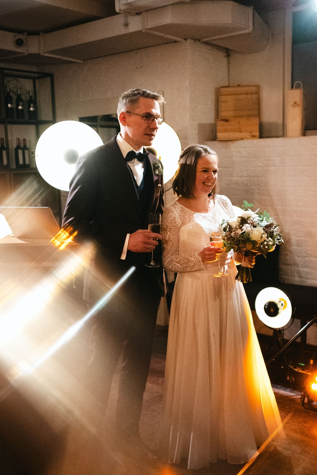 Mężczyzna w smoking z butonierką i kobieta w jasnej sukni z bukietem kwiatów na tle dekoracji sali weselnej.