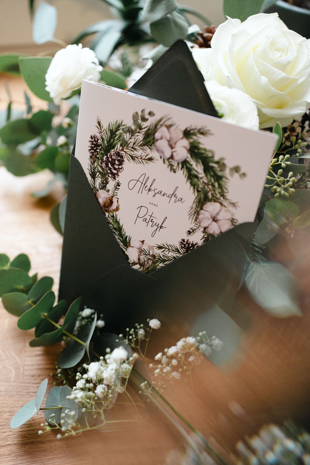 Papeteria ślubna z białymi różami, zaproszenie z personalizacją imion Alexandra i Patryk, otoczone zielenią i kwiatami na drewnianym stole