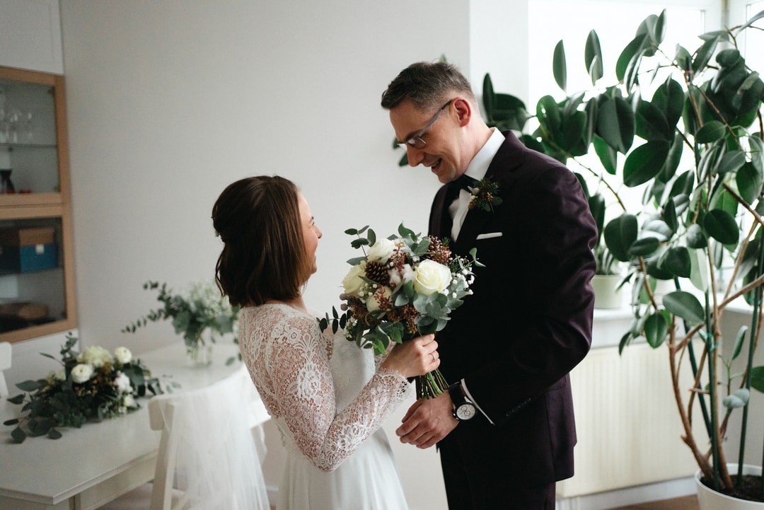 Para Młoda na bukiet ślubny podczas ceremonii w białej sali. Mężczyzna w czarnym garniturze z łękiem, kobieta w koronkowej sukni ślubnej. W tle rośliny doniczkowe i dekoracje florystyczne.