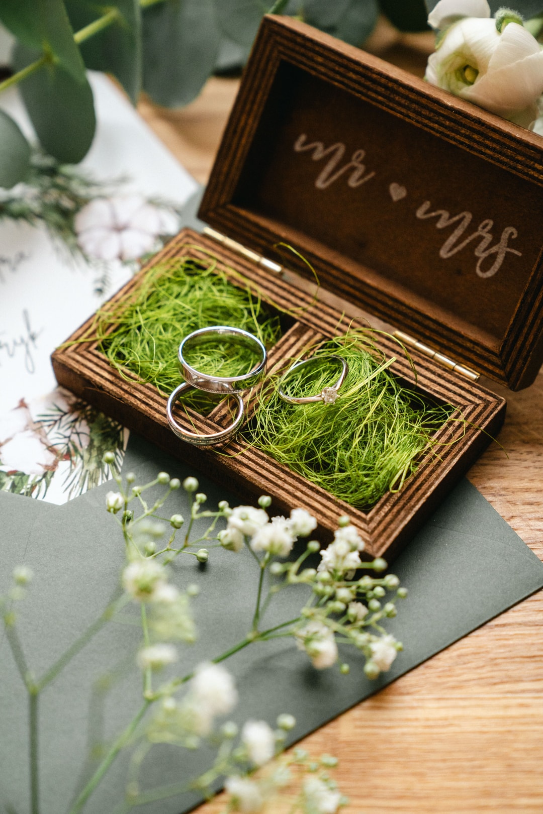 Drewniane pudełko na obrączki ślubne z napisem, wypełnione zielenią, dwie srebrne obrączki, białe kwiaty w tle