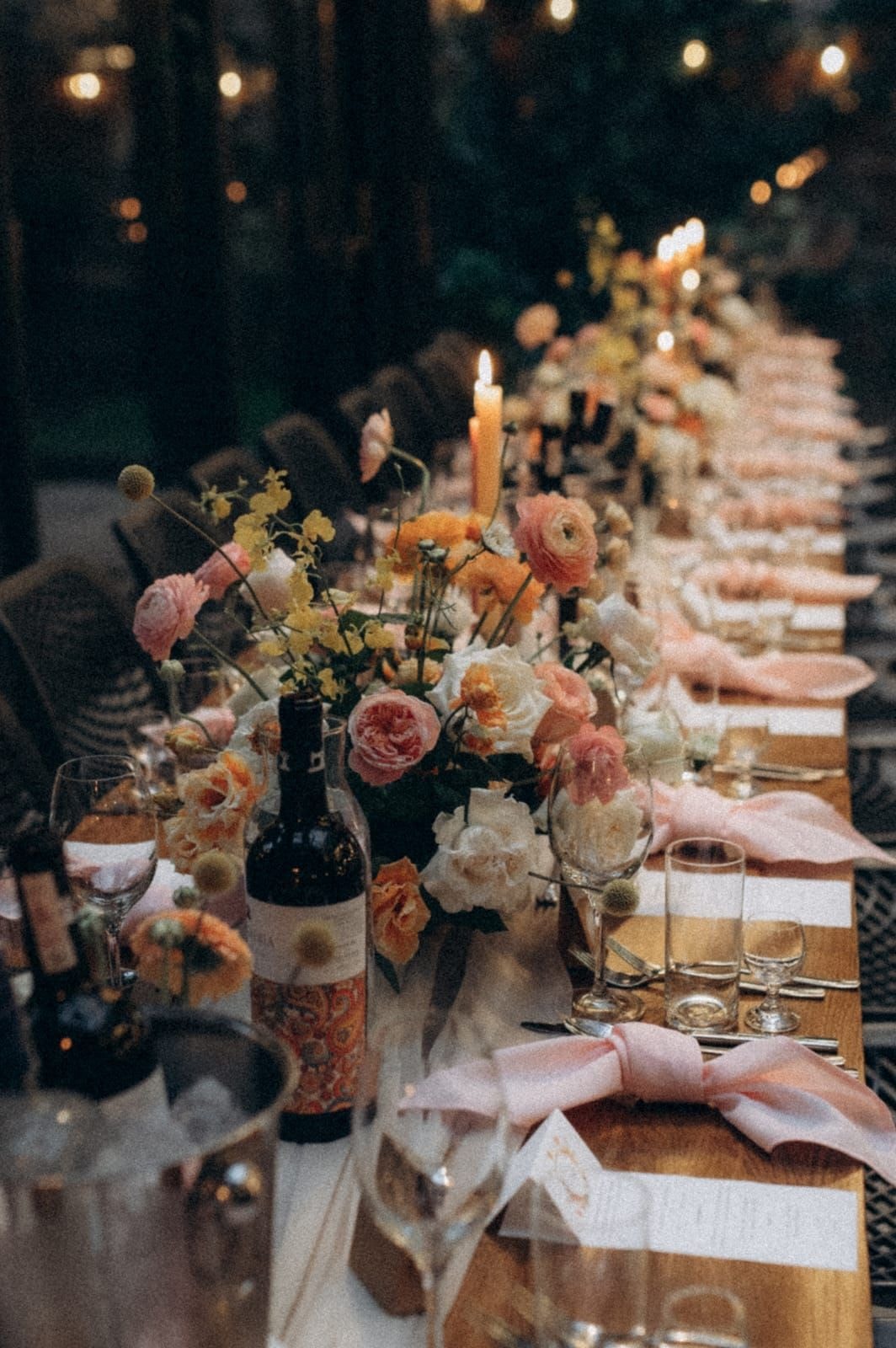 Długi stół z aranżacją stołów weselnych, kwiaty w pastelowych tonach, butelka wina, świece, goście w tle