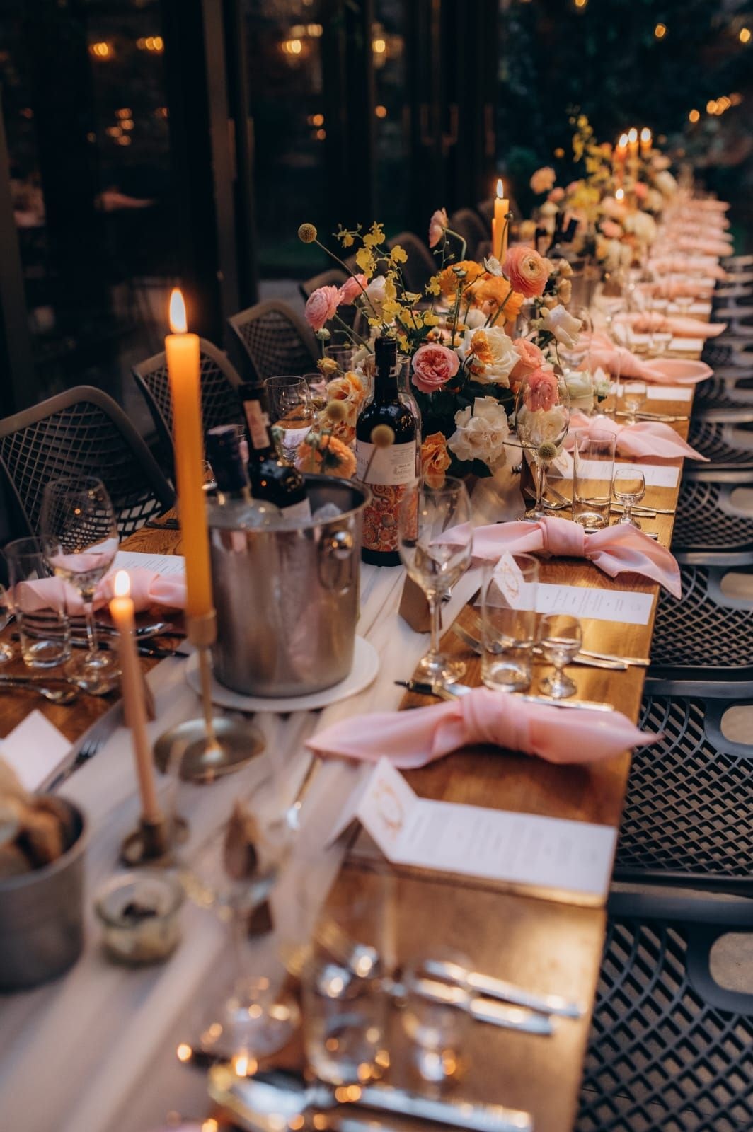 Długi stół z aranżacją kwiatową, świecami, kieliszkami i różowymi serzetkami na czarnych krzesłach