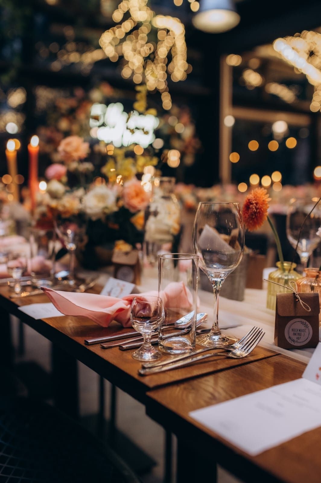 Aranżacja stołu weselnego z kieliszkami, różowymi serwetkami, sztućcami i kwiatami w tle, oświetlenie ze złotymi lampkami