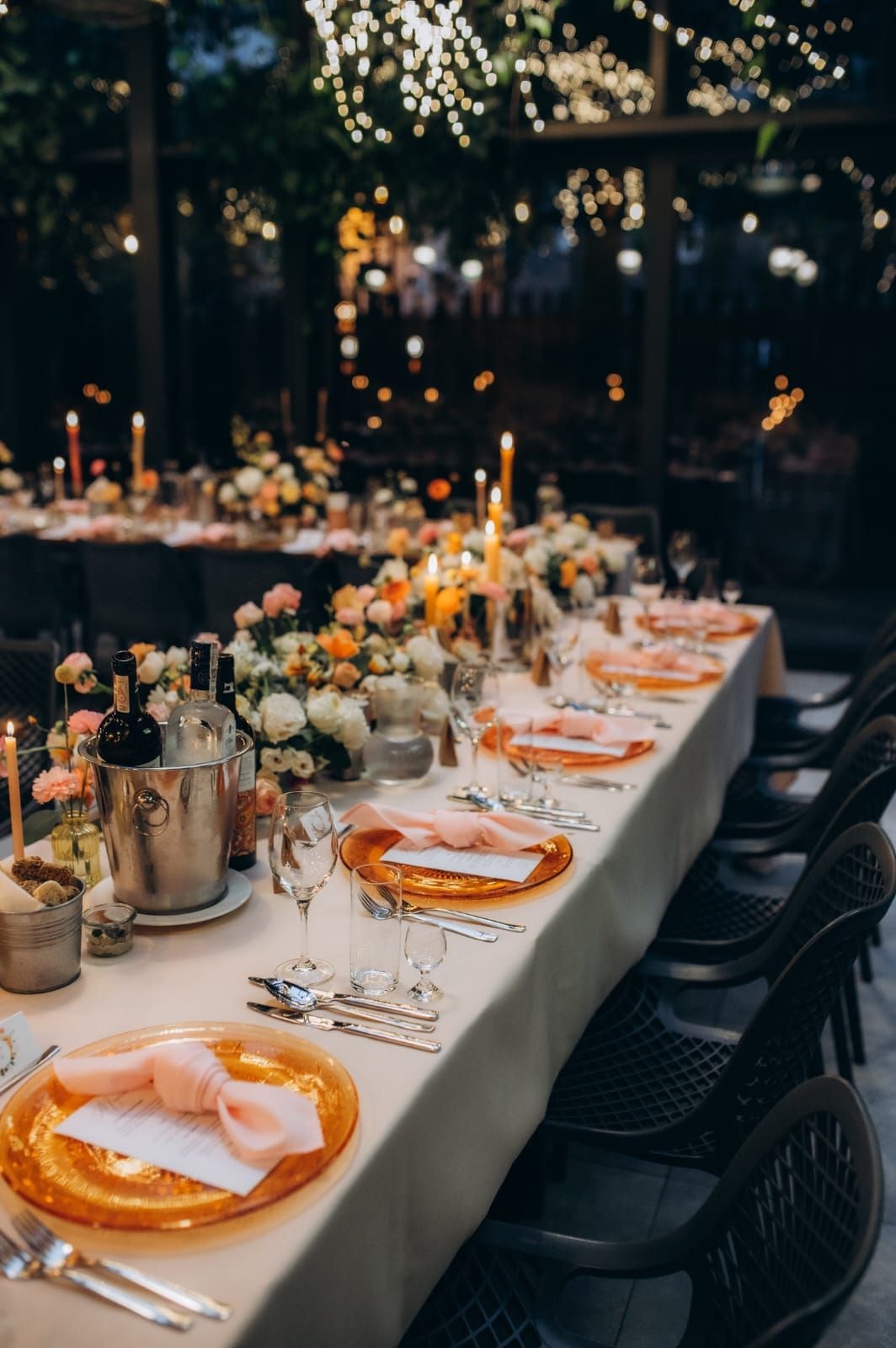 Długi stół weselny z białym obrusem, dekoracja stołu weselnego z kwiatami w jasnych tonach, złotymi świecznikami i pomarańczowymi talerzami