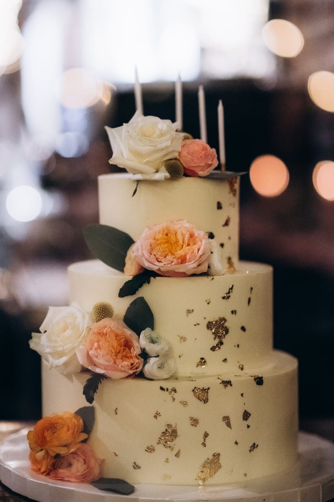 Trzypiętrowy tort weselny z białym lukrem, złotymi akcentami i świeżymi kwiatami w jasnej palecie na każdym piętrze, zdobiony topperami.