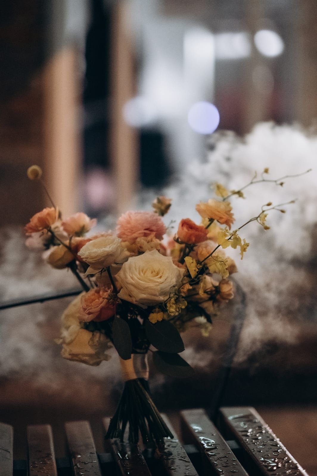 Bukiet ślubny z kremową różą i kwiatami w tonacji łososiowej, z delikatną zielenią, na drewnianym stole