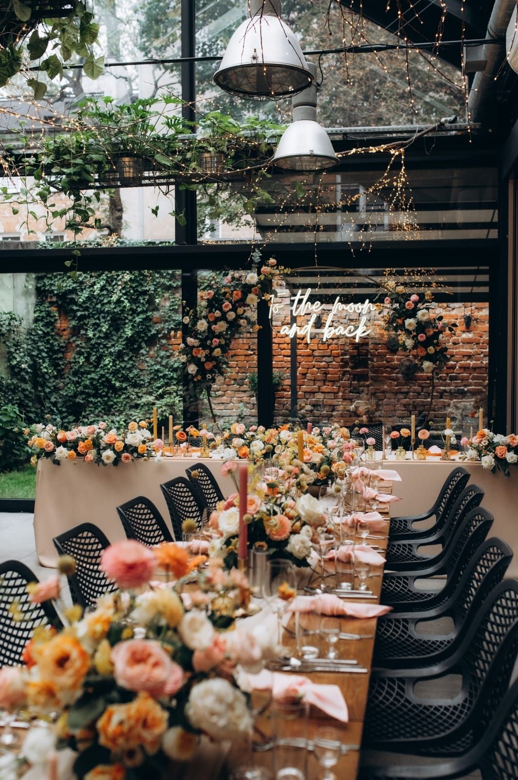 Aranżacja stołów weselnych z dekoracją kwiatową w tonacji jasnych i pomarańczowych kwiatów, czarne krzesła, długi stół, nowoczesna sala z cegłą i szkłem