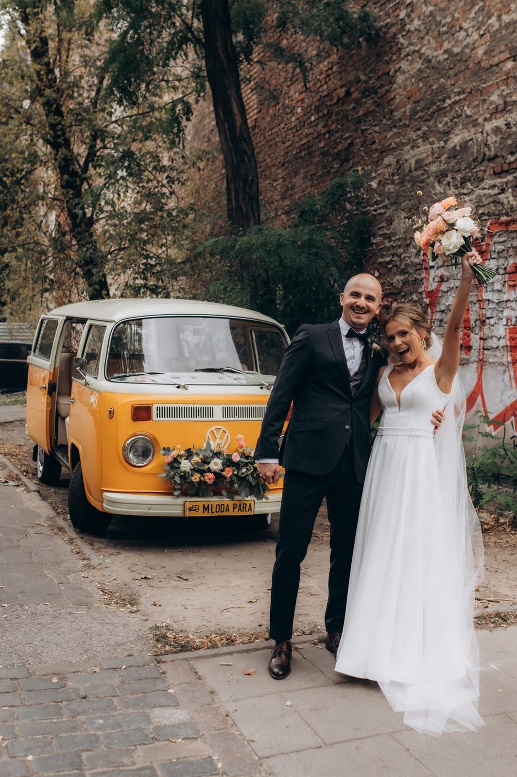 Para młoda przy żółtym autobusie z bukietem kwiatów w jasnej palecie, złocisty samochód ozdobiony kwiatami i zielenią na tle starej ceglanej ściany