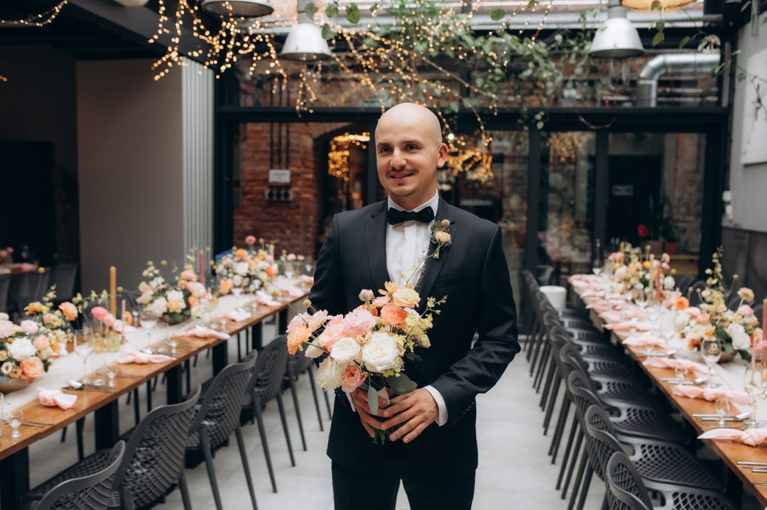 Mężczyzna w czarnym smokingu z muszką trzyma bukiet ślubny z białymi i łososiowymi kwiatami w elegancko udekorowanej sali weselnej.