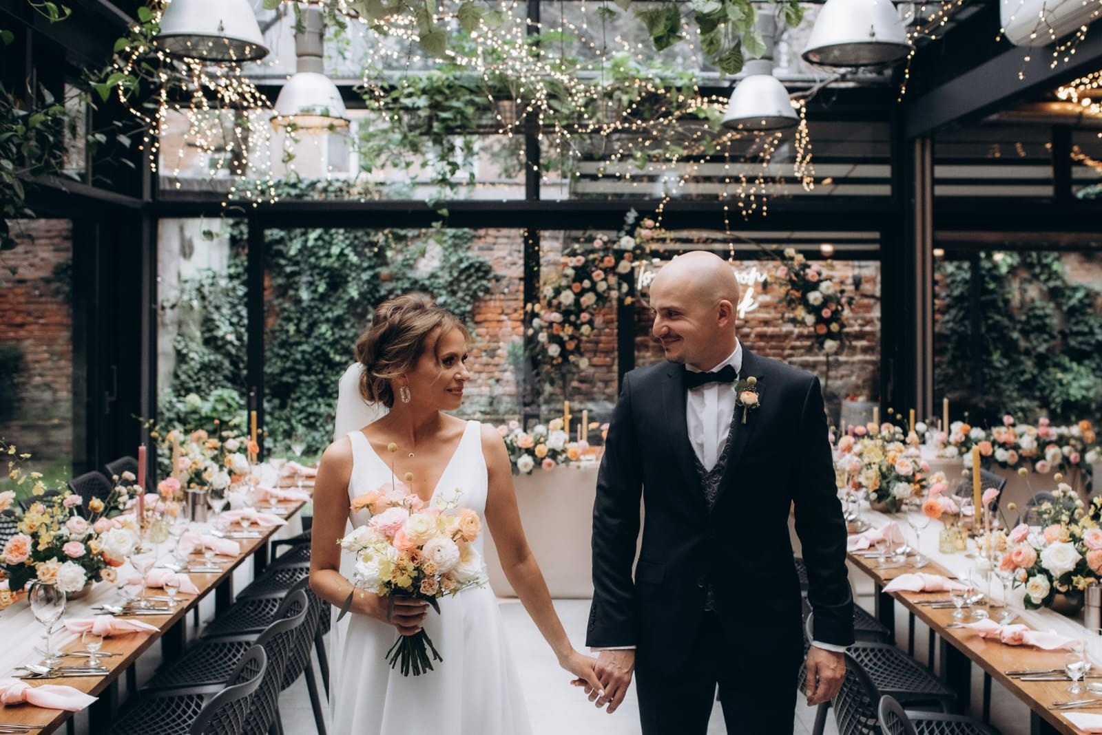 Para Młoda spaceruje między stołami na dekoracje sali weselnej z jasnych kwiatów, zieleni i świecącymi girlandami w ceglastej sali.