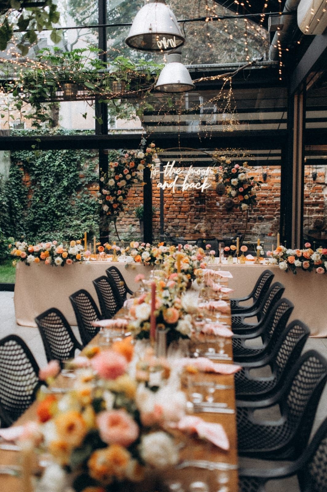 Aranżacja stołów weselnych z długim stołem, czarnymi krzesłami, dekoracjami z kwiatów w jasnej palecie i białymi lampami nad halą z ceglaną ścianą