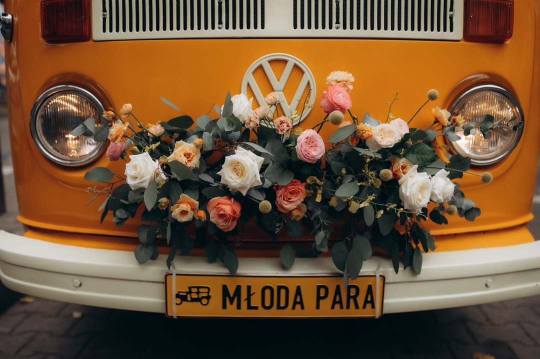 Dekoracja kwiatowa na przód pojazdu z białymi i łososiowymi kwiatami, zielenią i drewnianym logiem marki, tablica rejestracyjna Młoda Para