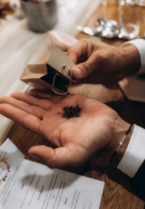 Dłonie mężczyzny trzymające papierowe pudełko z czarnymi nasionami, papeteria ślubna na drewnianym stole