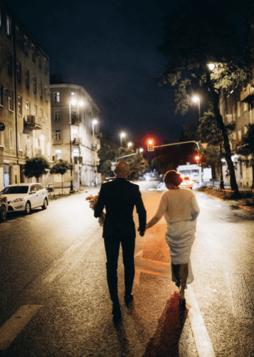 Dwoje ludzi spacerujących nocną ulicą wśród oświetlonych budynków i drzew, w tle świetlne tła miasta.