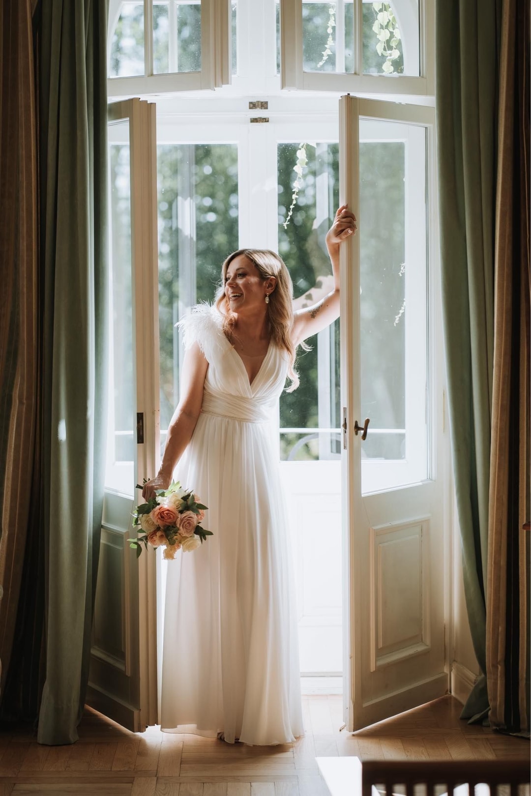 Panna młoda w białej sukni z bukietem kwiatów przy otwartych drzwiach, patrząca w ogród