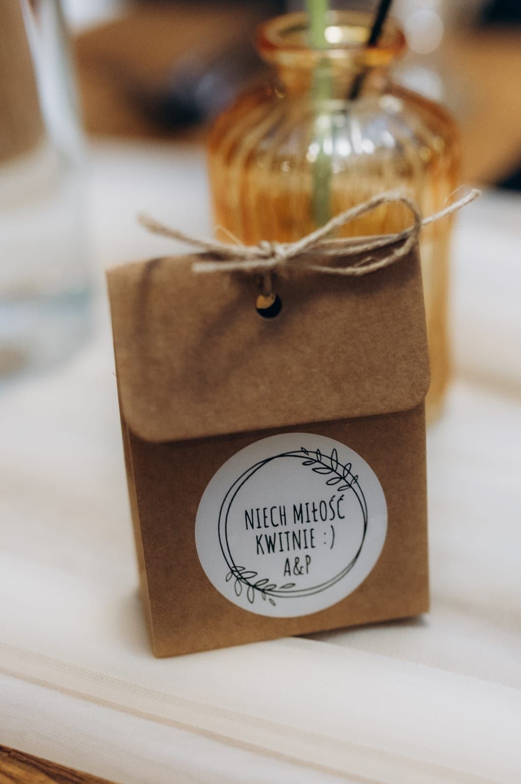 Papeteria ślubna: kraft'owy woreczek z białą etykietą, napisem "Niech miłość kwitnie" i inicjałami A&P, zawiązany sznurkiem, na białym blacie obok butelki perfum.